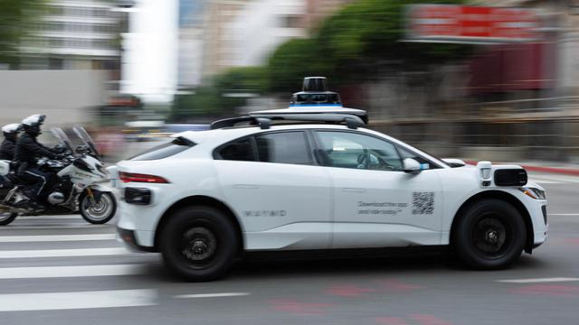 Police officers riding motorcycles follow a Waymo driverless vehicle as it drives through downtown Los Angeles